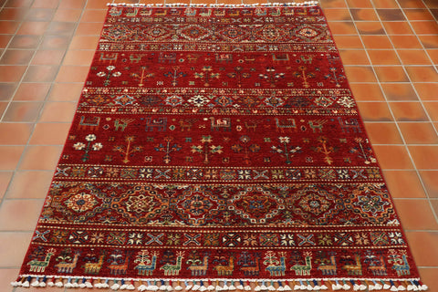 A rich red ground for all of this rug.  There are bands of decoration upon it showing various depictions of birds, animals and stylized floral designs.  The colours used to show these are a mix of greens, blues, oranges, lighter reds, golds and creams.  The bands of decoration are laid out symmetrically across the rug, alternating from animal/bird to flora or fauna. 