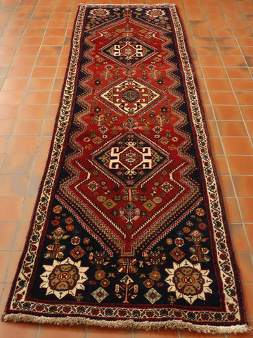 Warm red central ground with three decorated lozenges upon it.  The central one is cream, the other two navy blue.  At either end of the main section there is a broad area that is sectioned into 4 triangles using very dark blue.  Stylized floral decoration can be found in this area, using cream as the main colour and reds, olive greens and oranges to highlight.  Around the main flower are smaller ones.  There is a cream border with floral intertwined decoration using the same colour palette.  