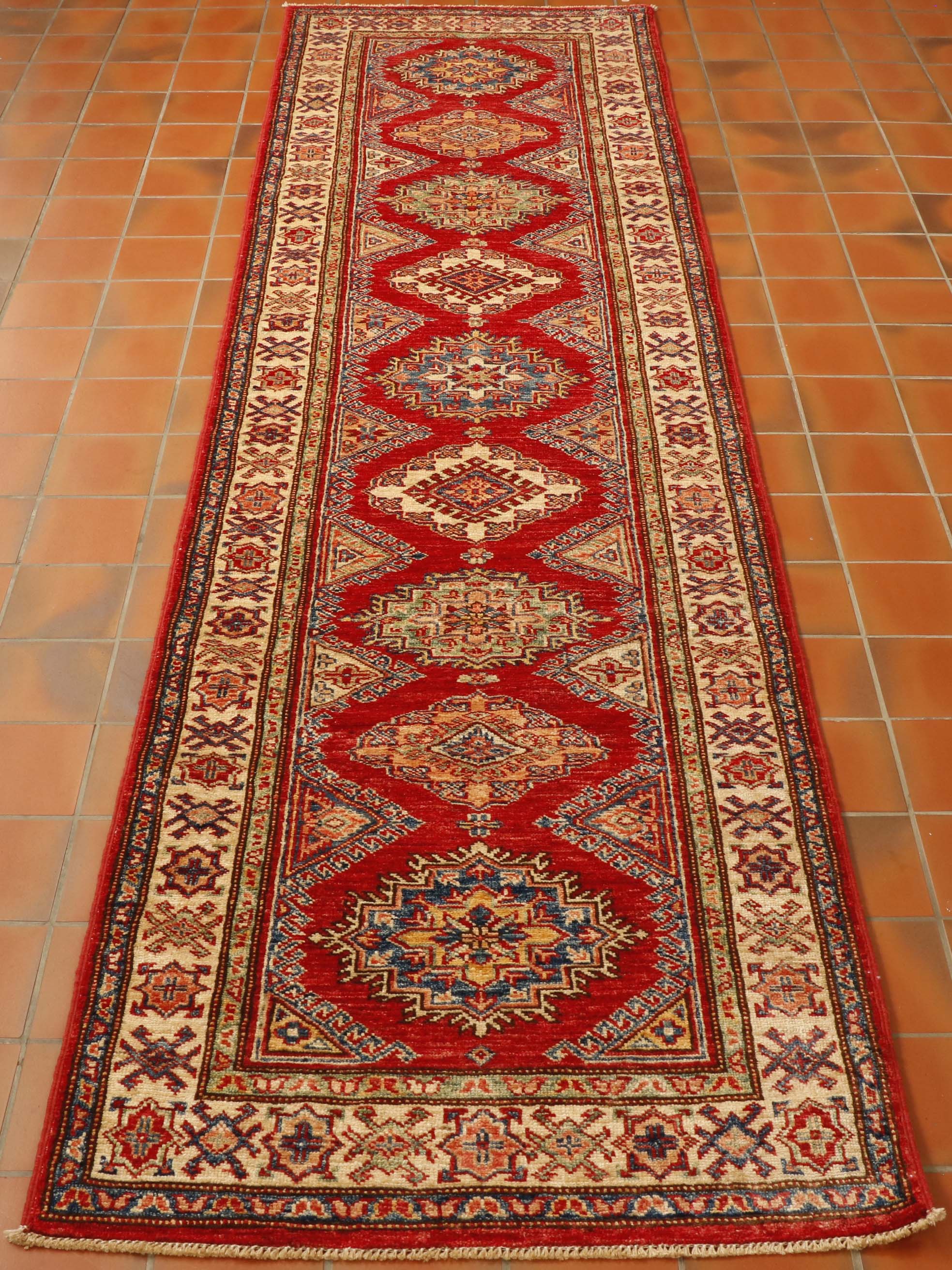 Bright red central ground with a series of medallions through nthe length.  Border in cream with narrower borders on either side.  Colours used in the decoration consist of dark blue, green, golden yellow, peach and cream. 